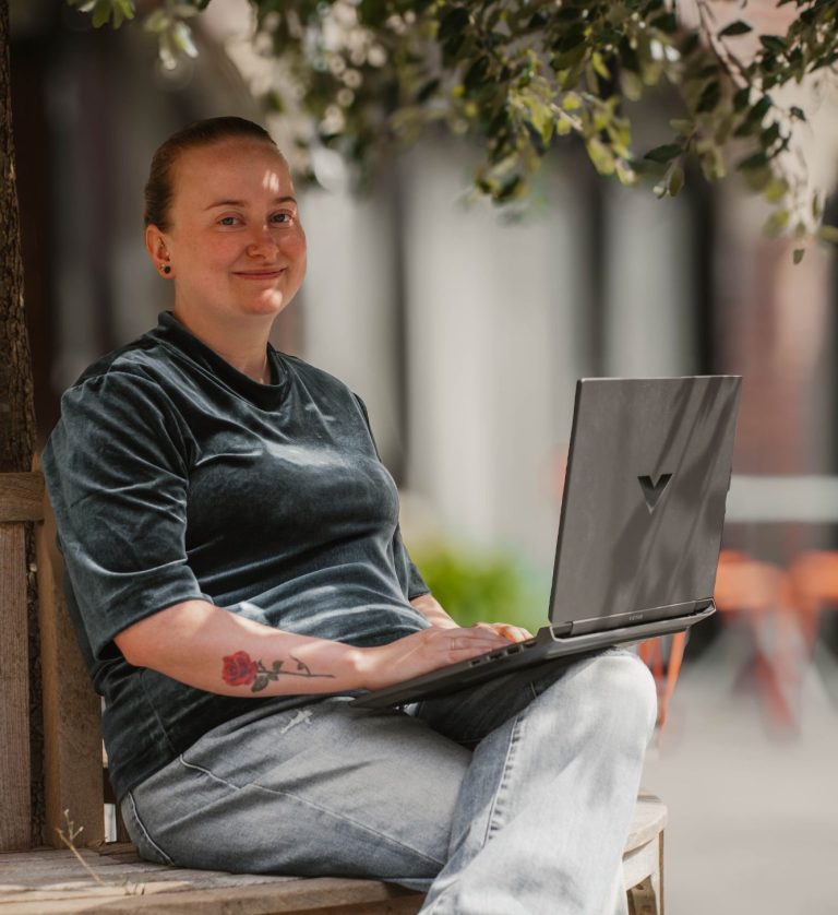 Taakmevrouw met een laptop, zittend op een bank in een groene, urbane omgeving.