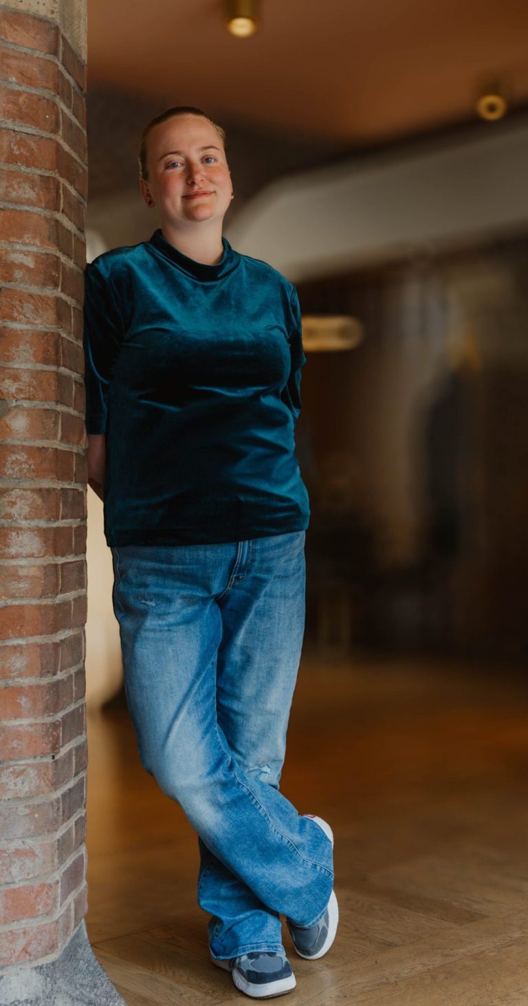 Taakmevrouw leunt tegen een muur in een lichte, moderne ruimte.