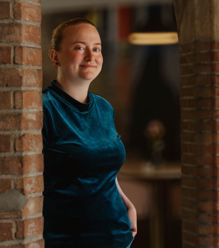 Taakmevrouw met een glimlach, gekleed in een blauwe top, leunt tegen een stenen pilaar.