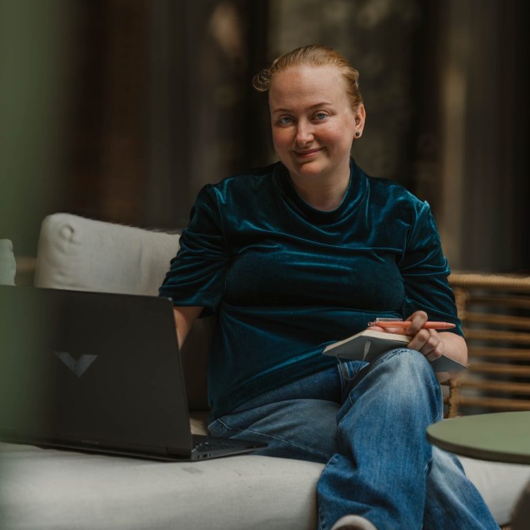 Taakmevrouw met een notitieblok en laptop, glimlachend op een bank in een moderne ruimte.